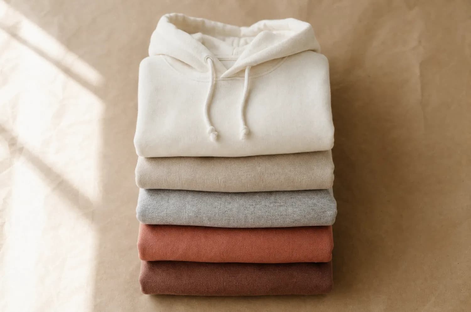 A stack of folded plain hoodies in cream, oat and clay tones.